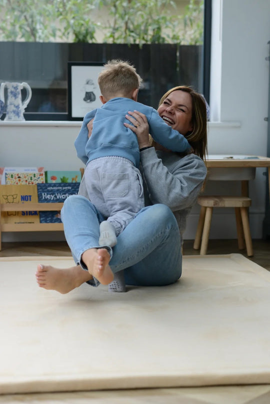 Mum laughing and lifting her young son on a cream plush Momomi play mat in a bright playroom