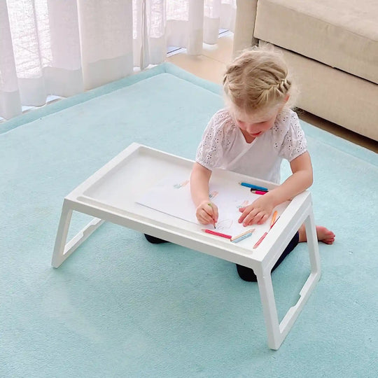 Young girl kneeling on a mint carpet, drawing with coloured pencils at a white foldable kids table