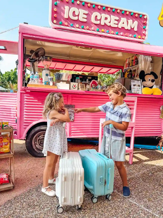 Two children with white and blue Momomi kids suitcases in front of a pink ice cream truck