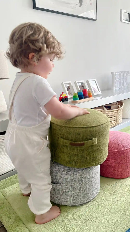 Toddler in white overalls stacking a green and grey Momomi kids pouffe chair on a play mat at home