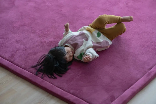 Toddler lying back and relaxing on a soft berry plush Momomi nursery rug over a wood floor