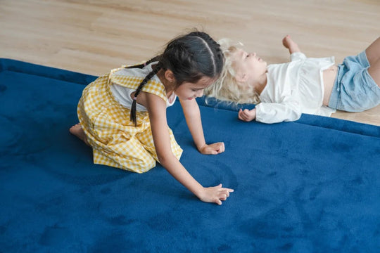 Two young girls playing and relaxing on a large navy foam play mat on a wooden floor