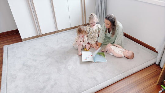 Woman and two toddlers reading on gray plush play mat in bright modern living room with wooden floors